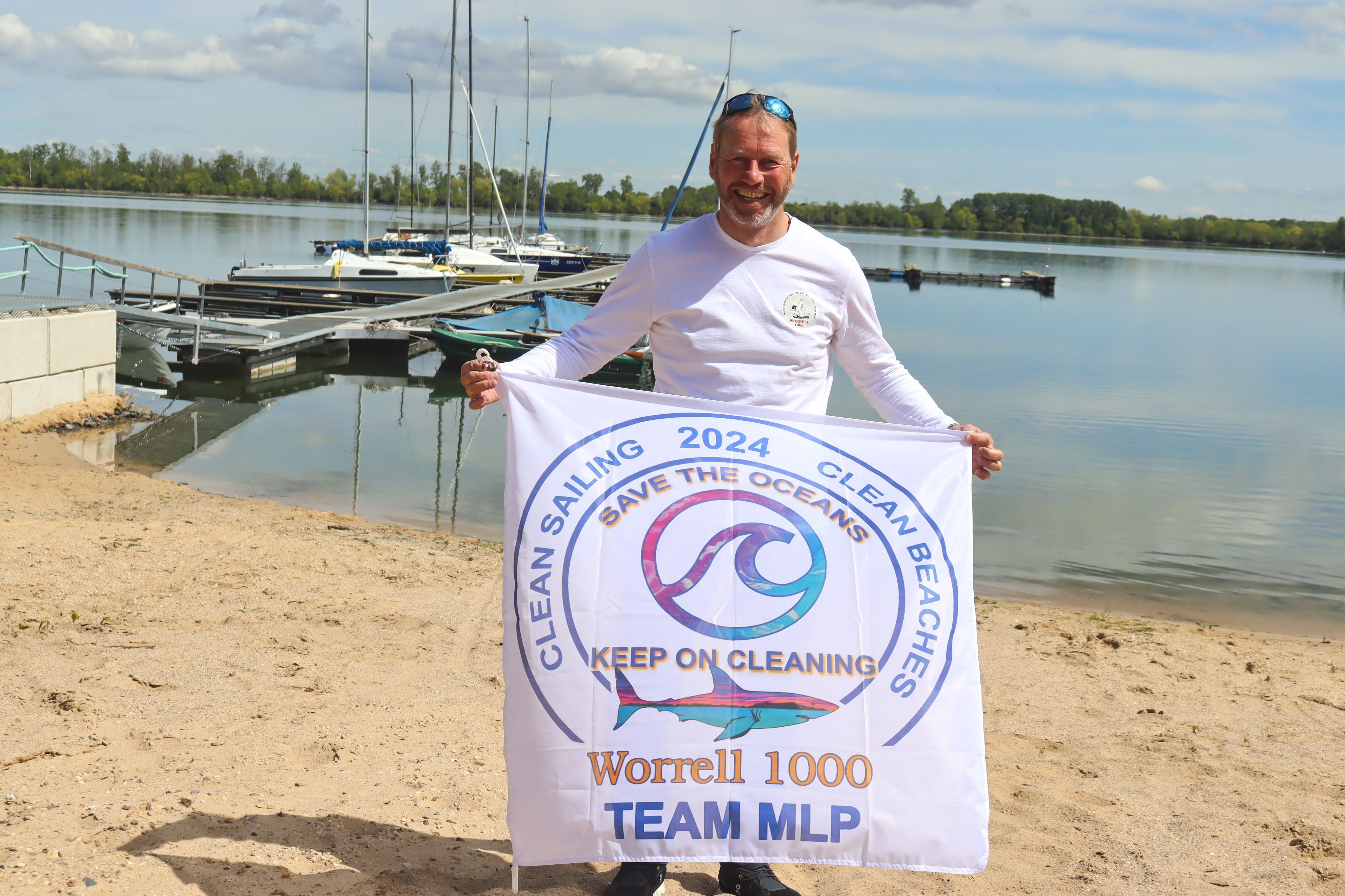 André Hauschke hält eine Fahne hoch, auf der „Clean Sailing 2024 Clean Beaches, Save the Oceans, Keep on Cleaning“ und der Hinweis „Worrell 1000 - Team MLP“ stehen. Im Hintergrund sind auf einem See Segelboote zu erkennen.