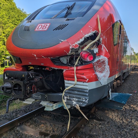 Eine Bahn, die an ihrer Front beschädigt wurde, steht auf Schienen. In der Umgebung stehen Bäume.