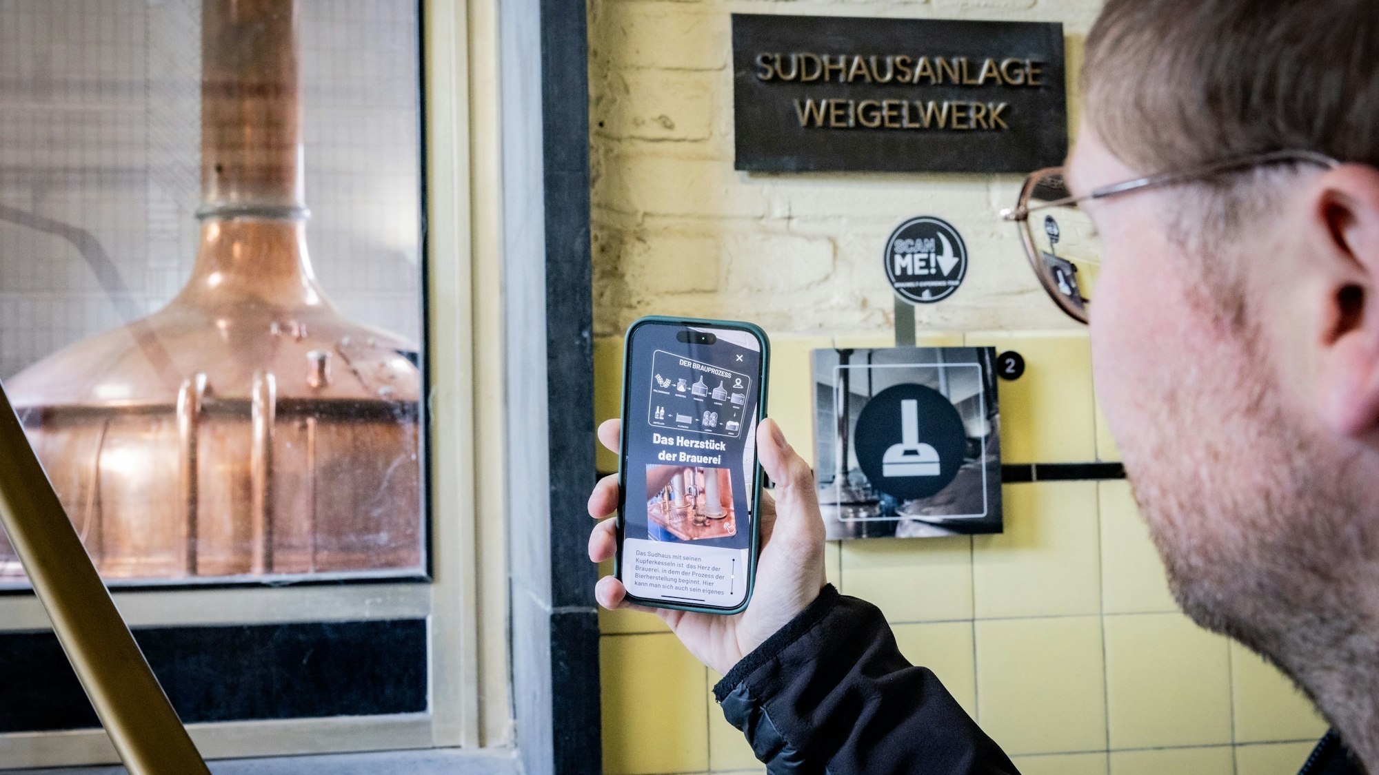 Ein Mann scannt ein Schild. Im Hintergrund steht einen großen Messingbottich in der Brauerei.