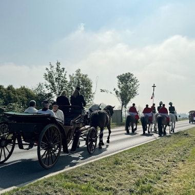 Zu sehen sind Reiter und Kutschen, die auf einer Straße unterwegs sind.