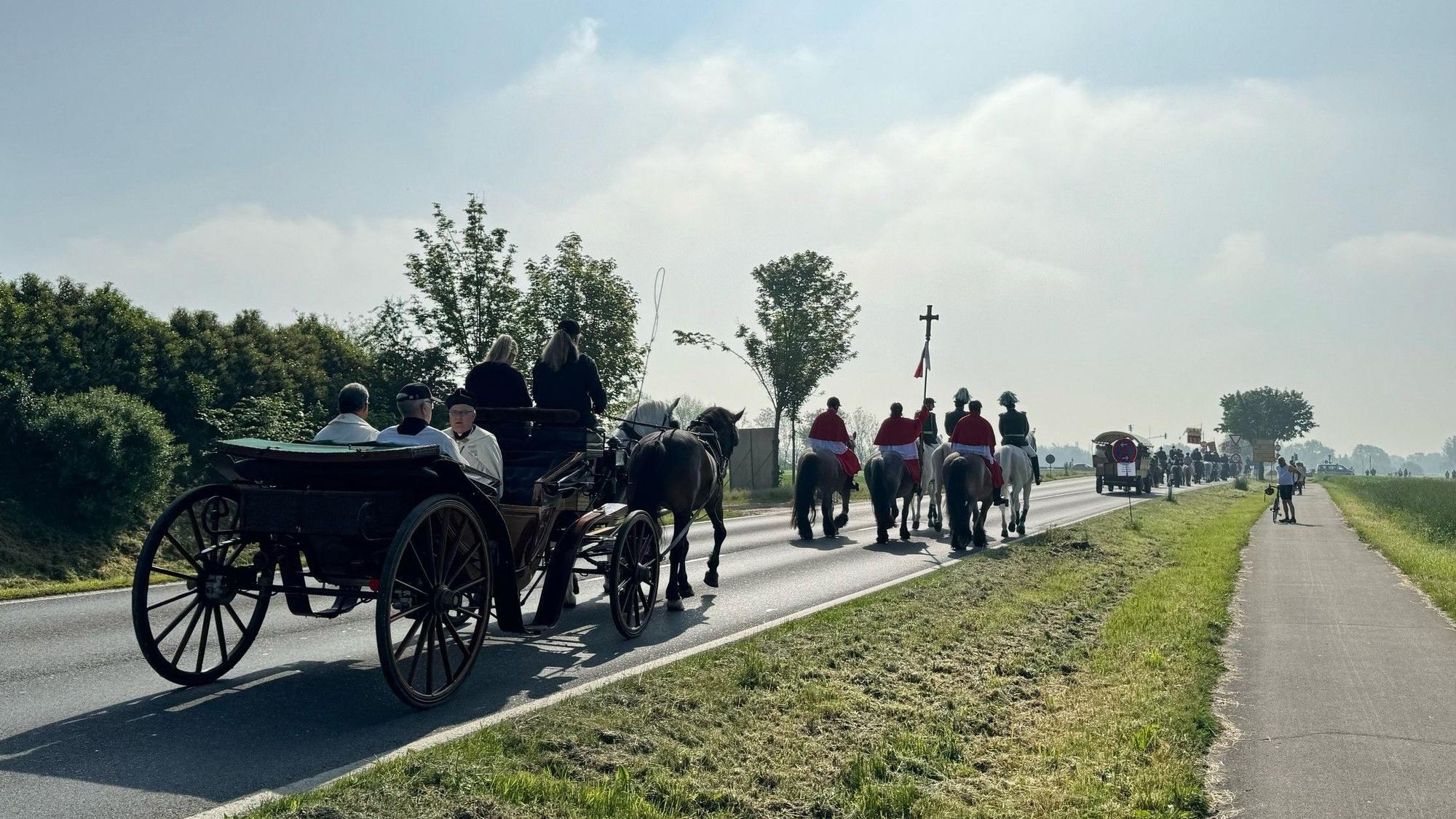 Zu sehen sind Reiter und Kutschen, die auf einer Straße unterwegs sind.