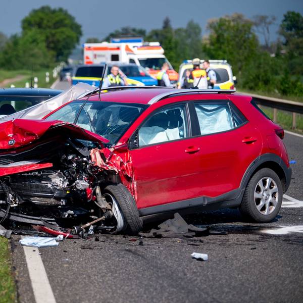 Ein zerstörter roter Kleinwagen steht auf einer Straße bei Euskirchen. Die Fahrerin ist bei dem Unfall ums Leben gekommen. Im Hintergrund sind Polizeifahrzeuge und ein Rettungswagen zu sehen.