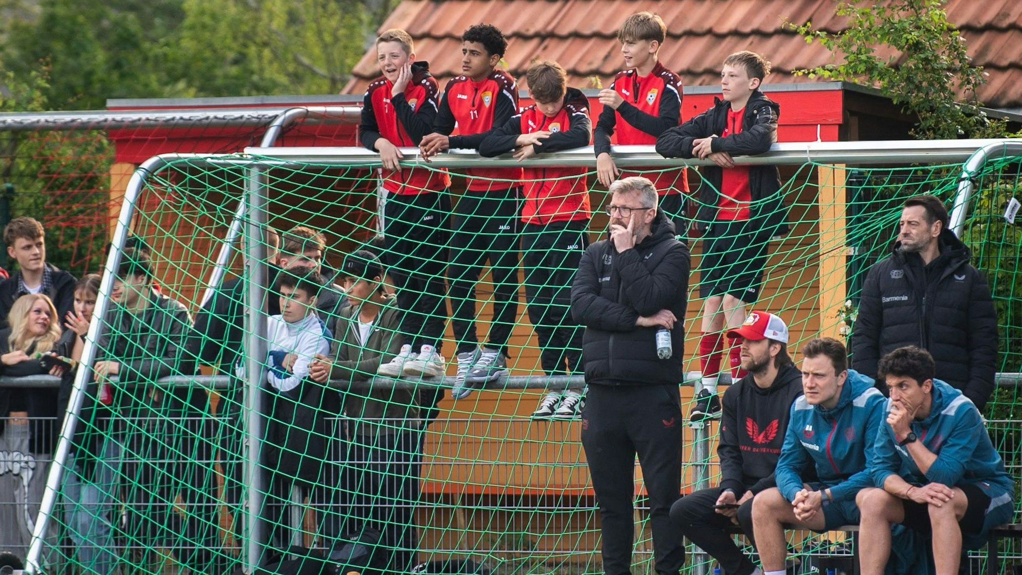 Fünf Jugendfußballer in schwarz-roten Trainingsanzügen stehen auf der Platzbegrenzung in Kuchenheim und halten sich an einem Tor fest, um das Spiel der A-Jugend gegen Bayer Leverkusen zu sehen.