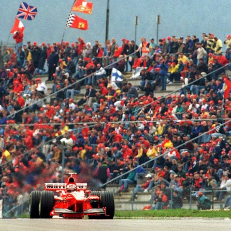 Der rote Rennwagen von Michael Schumacher fährt 1998 vor einer Tribüne voller Fans mit roten Käppis.