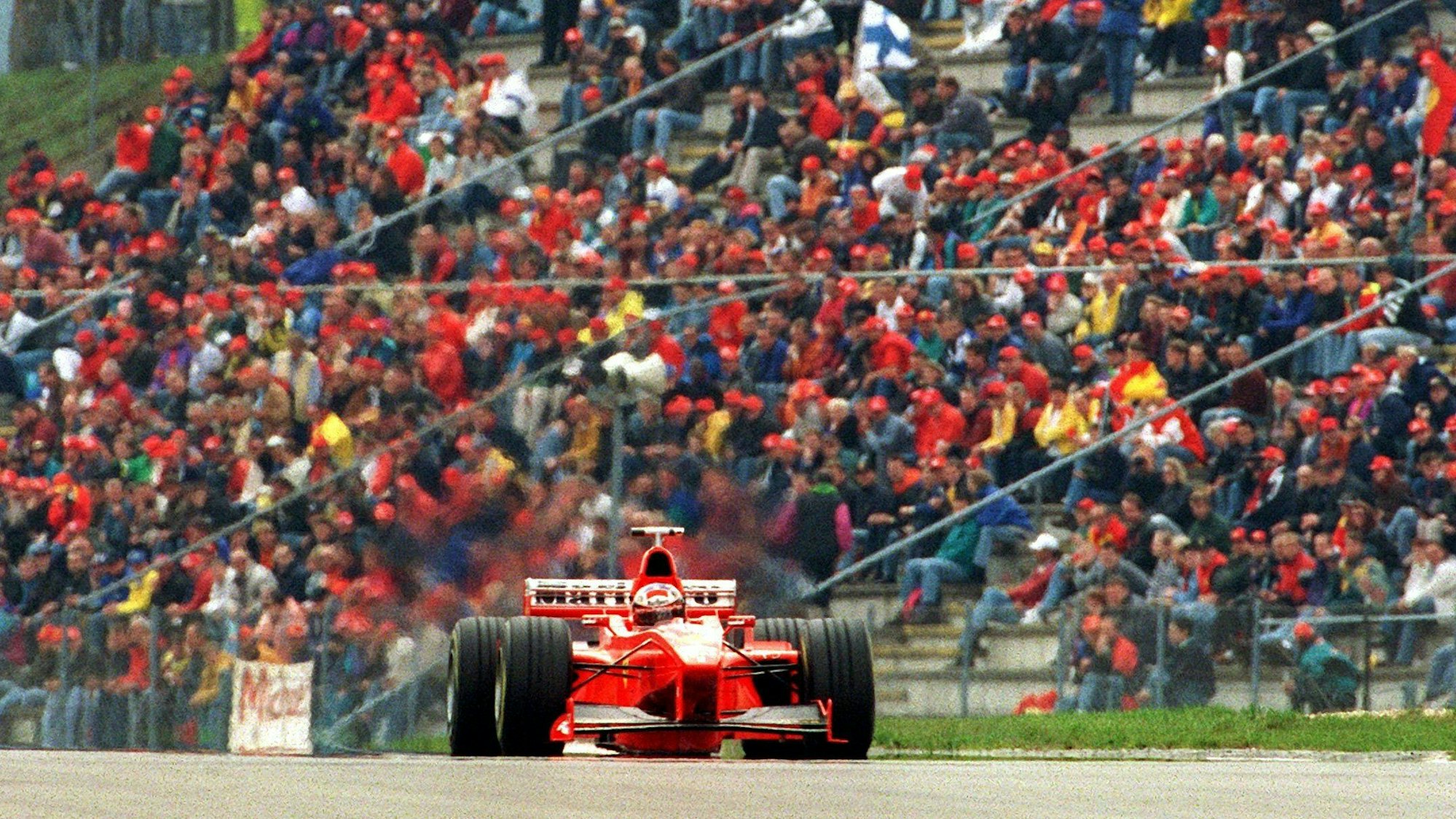 Der rote Rennwagen von Michael Schumacher fährt 1998 vor einer Tribüne voller Fans mit roten Käppis.