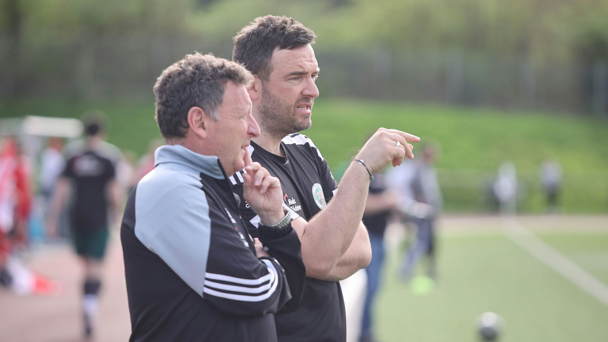 Zwei Trainer stehen am Rand eines Fußballfeldes nebeneinander.