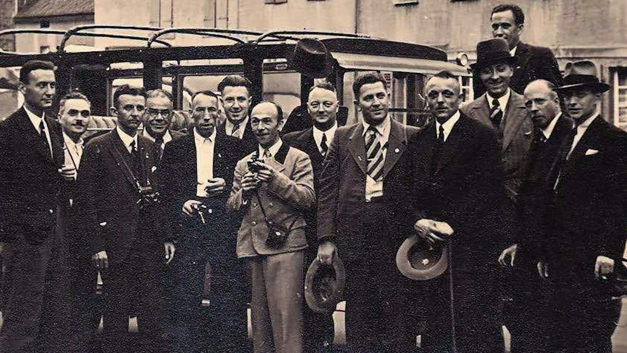 Auf einem historischen Bild stehen mehrere Männer vor einem Auto.