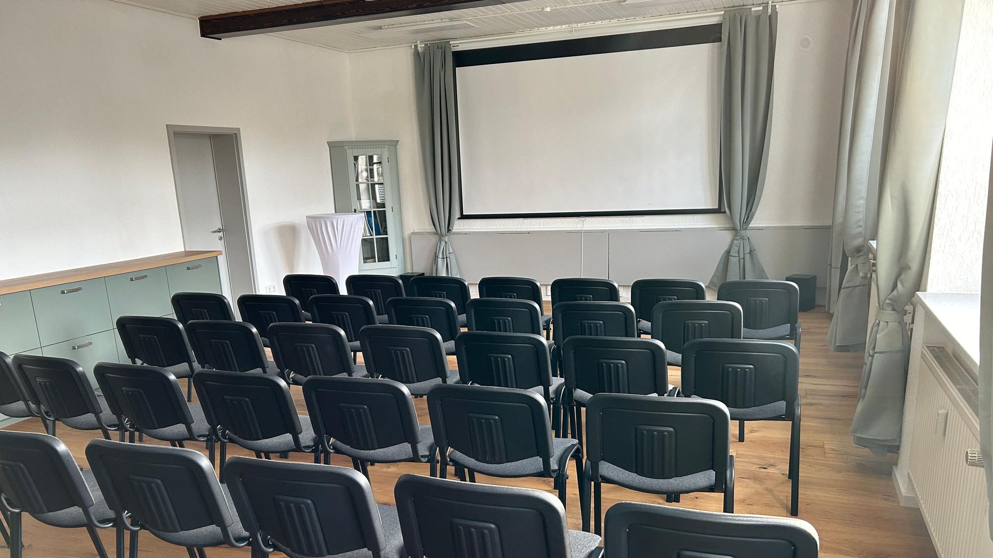 Im Saal stehen Stuhlreihen vor einer Leinwand.