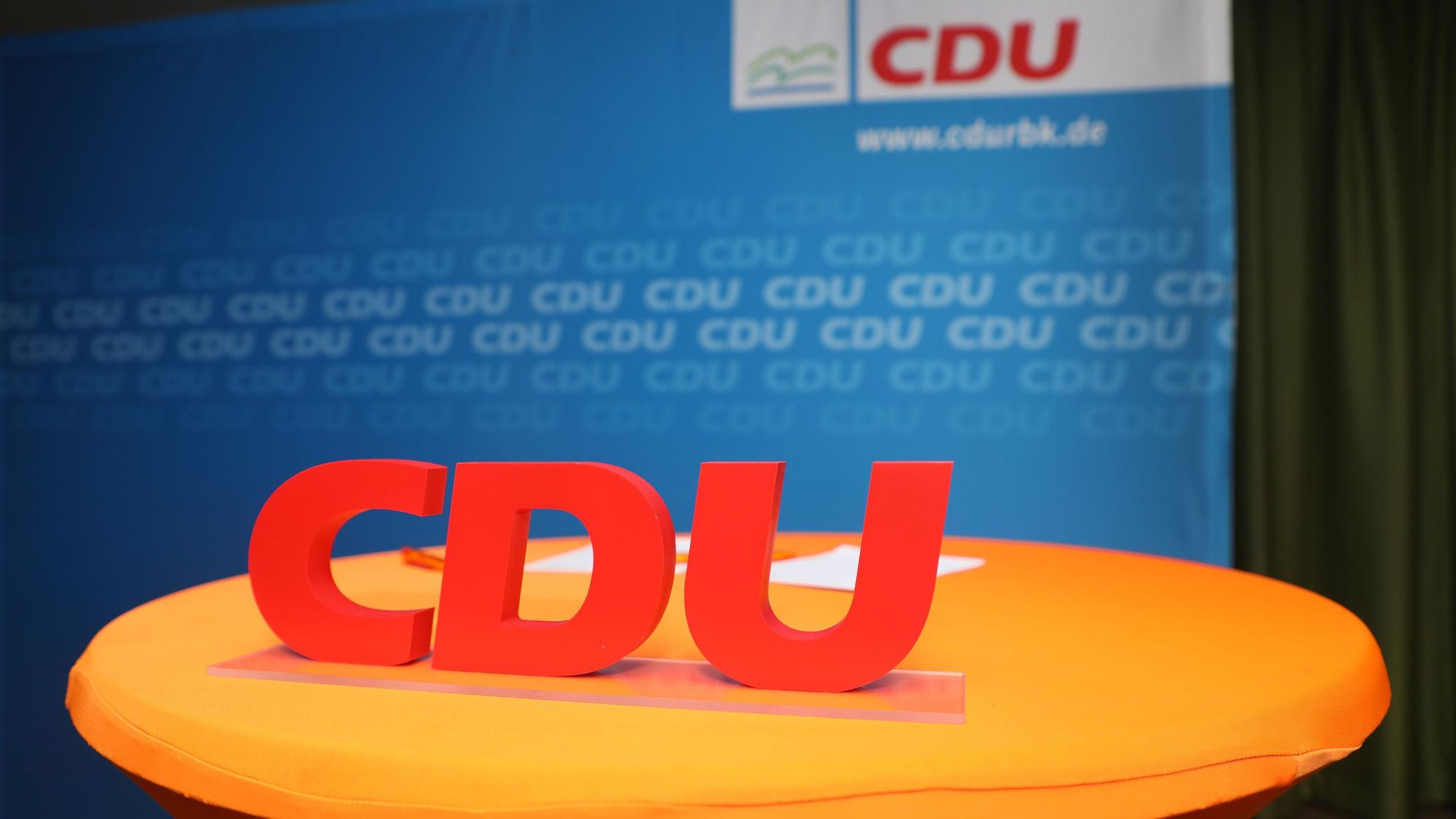 Vor einem blauen Plakat mit CDU-Aufschrift steht in großen roten Buchstaben CDU, platziert auf einem orangefarbenen Tisch.