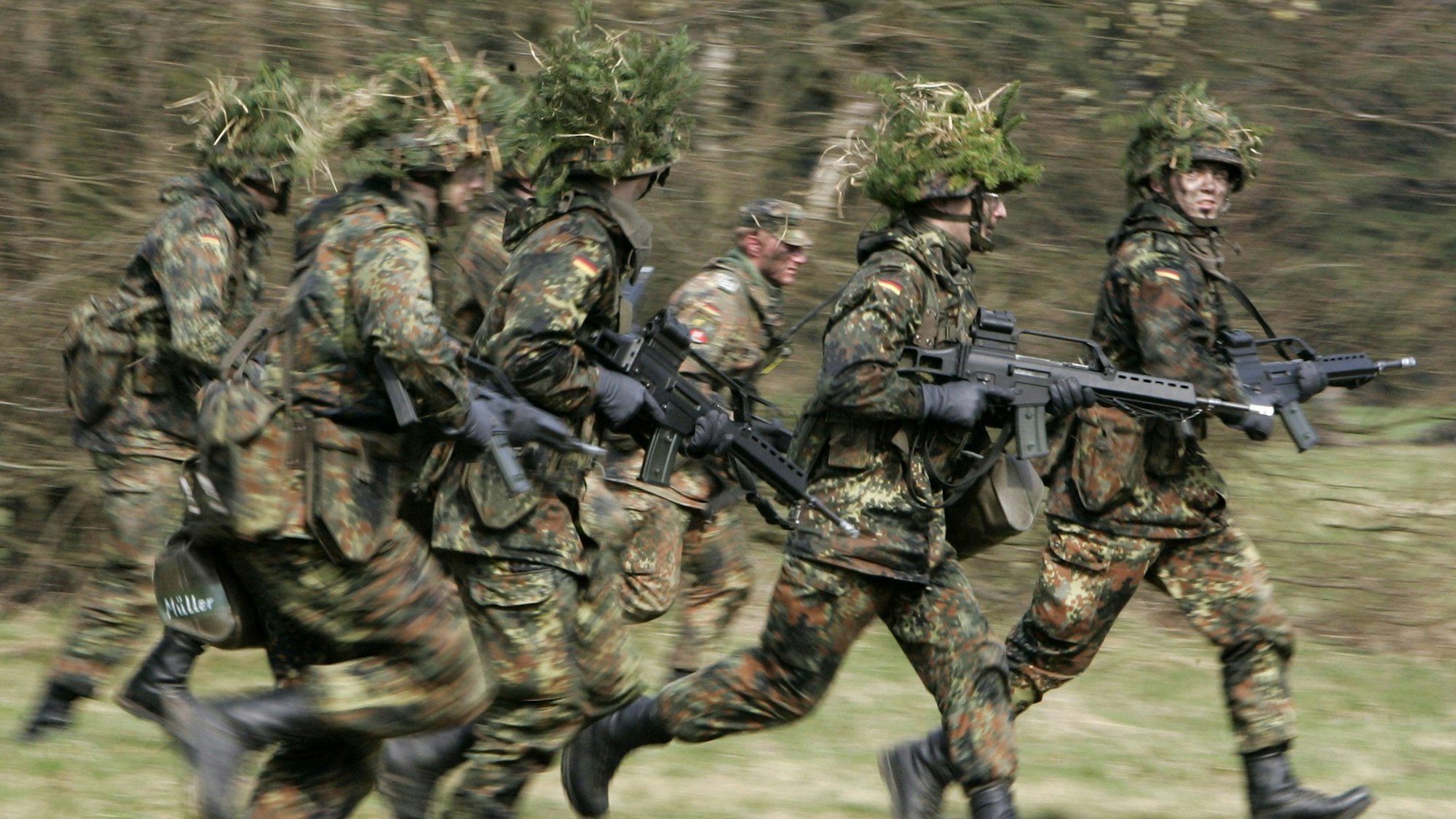 ARCHIV - 14.04.2005, Hessen, Schwarzenborn: Mit Blattwerk und Gras getarnte Wehrpflichtige von den Panzergrenadieren rennen mit ihren Waffen im Rahmen ihrer Grundausbildung bei einer Übung über das freie Gelände der Knüll Kaserne in Schwarzenborn (Schwalm-Eder-Kreis).