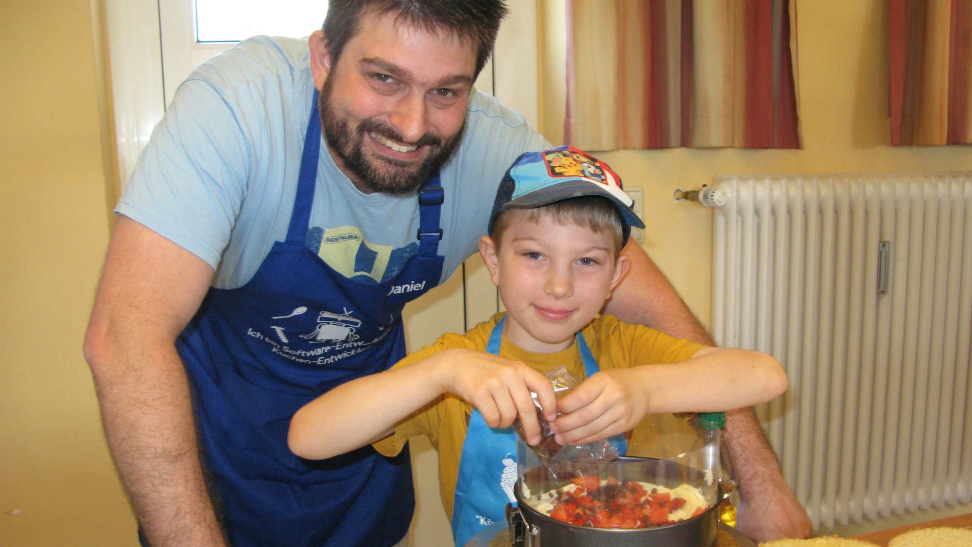 Ein Mann mit dunkelblauer Schürze und ein kleiner Junge mit Mütze beim Füllen der Torte mit Früchten.