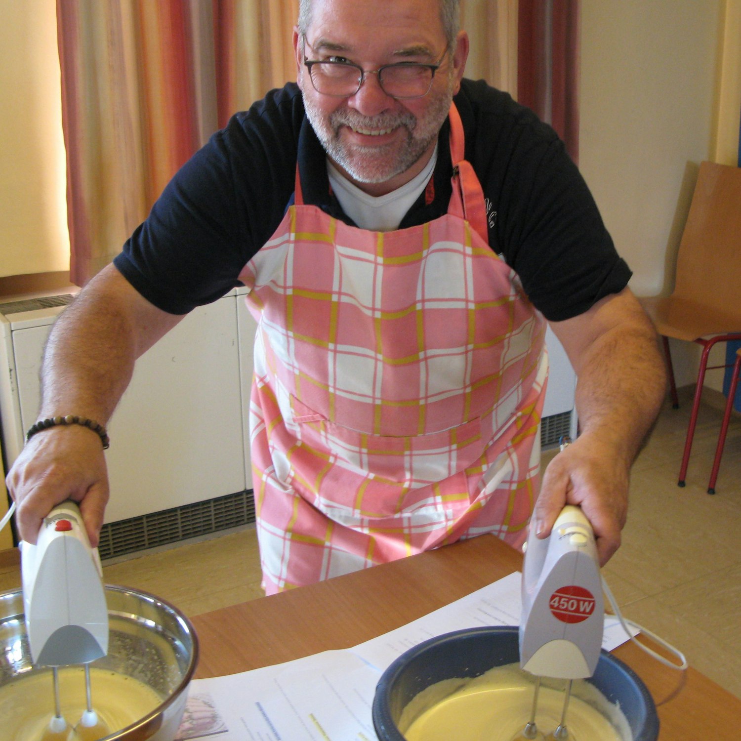 Ein Man mit karierter Küchenschürze mit zwei Handmixern in zwei Teigschüsseln.