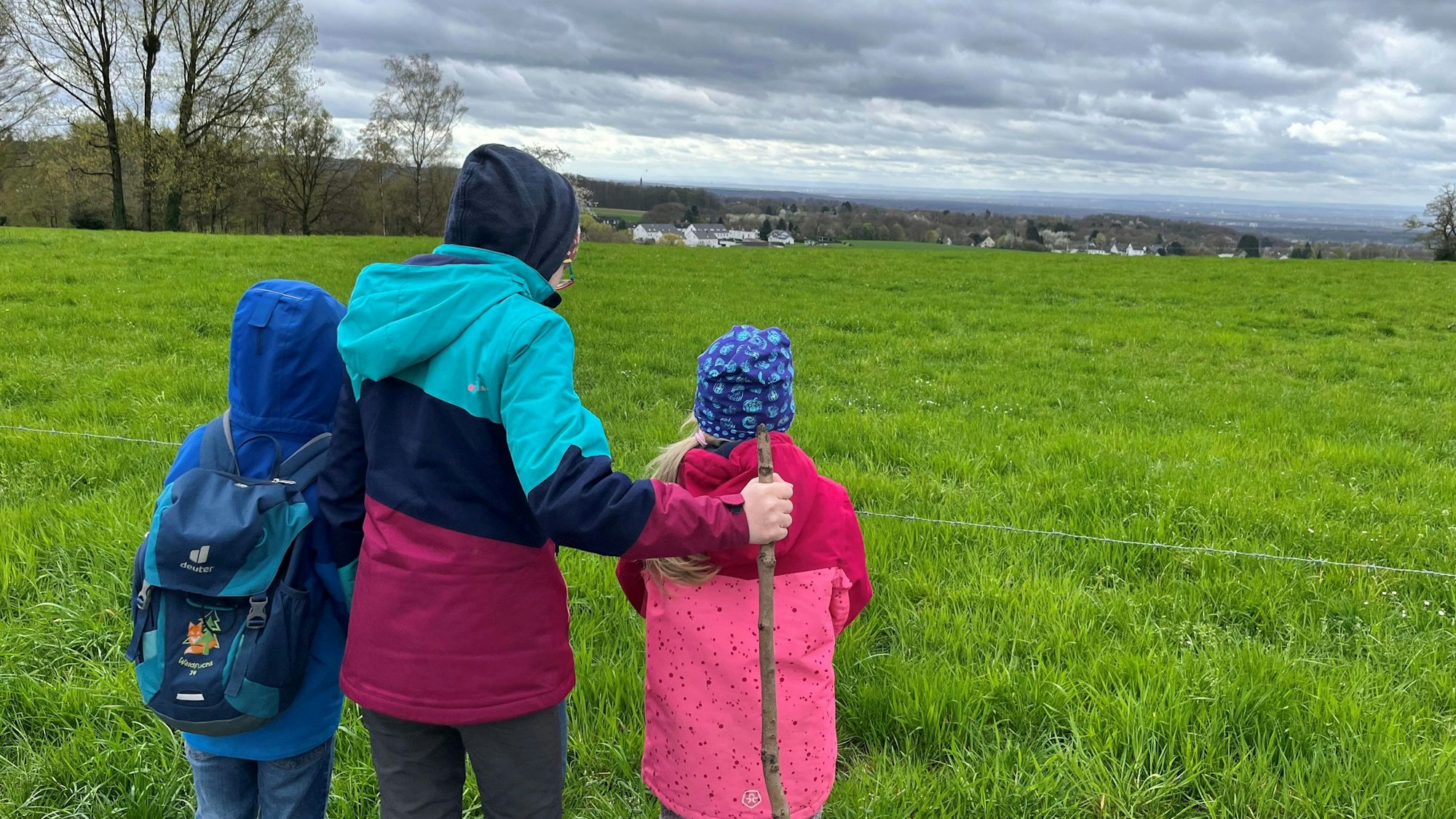 Drei Kinder stehen mit Mützen und Jacken vor einer Weide mit weitem Ausblick.