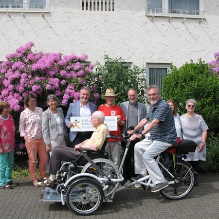 Die Rikscha mit Rollstuhlplattform, auf der ein Senior im Rollstuhl sitzt, fährt an eine Reihe von Menschen vorbei.