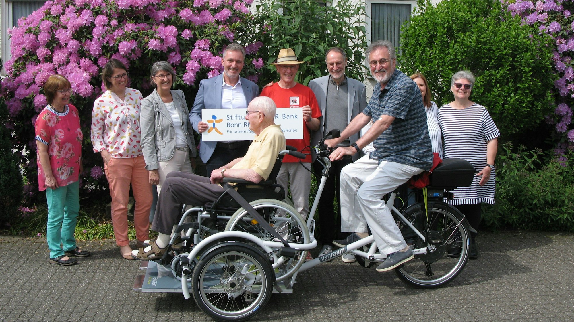 Die Rikscha mit Rollstuhlplattform, auf der ein Senior im Rollstuhl sitzt, fährt an eine Reihe von Menschen vorbei.