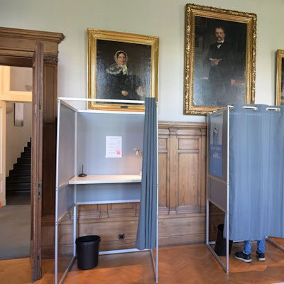 Drei Wahlkabinen stehen nebeneinander. In der mittleren Kabine sieht man die Füße eines Wählers.