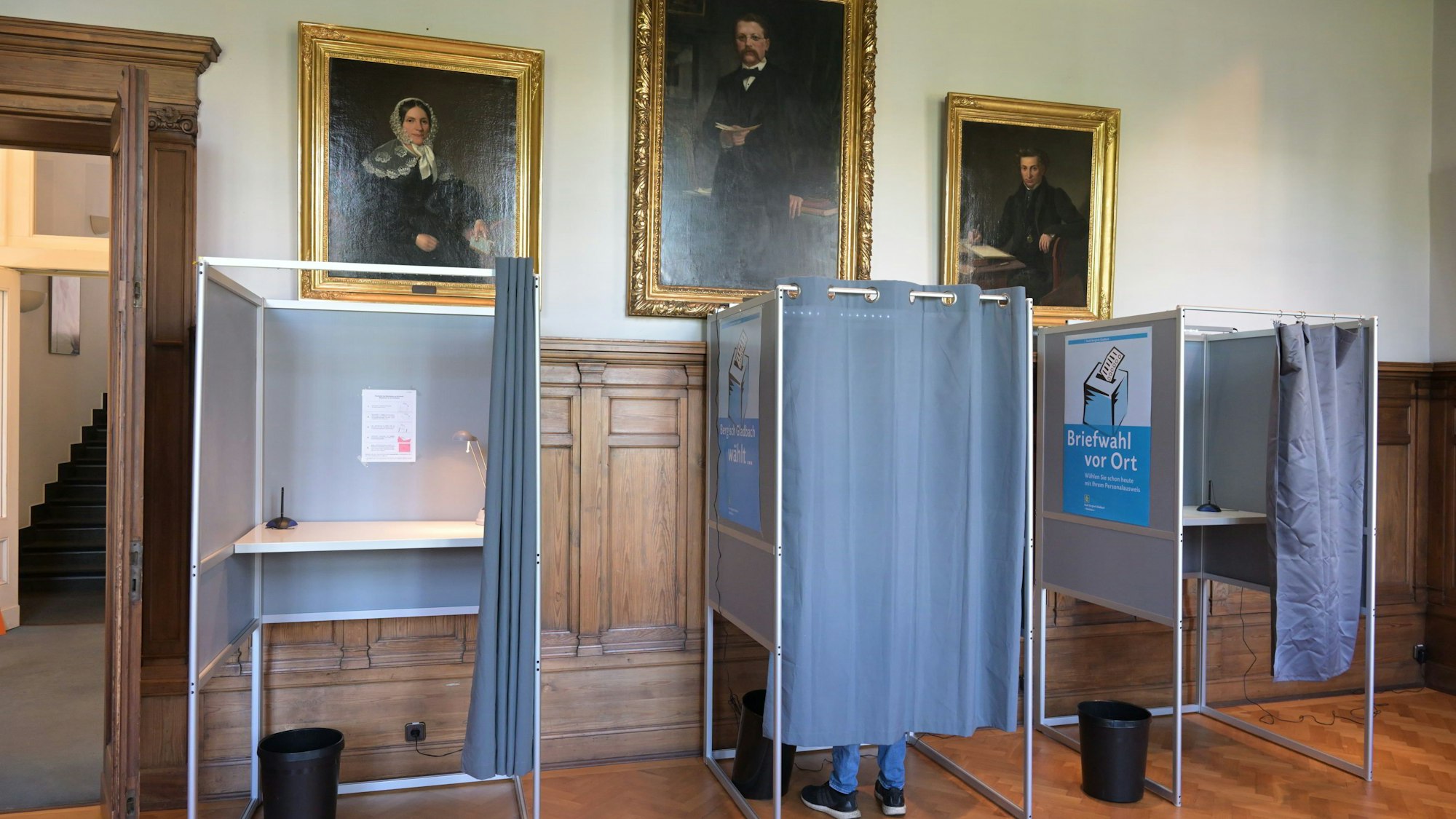 Drei Wahlkabinen stehen nebeneinander. In der mittleren Kabine sieht man die Füße eines Wählers.