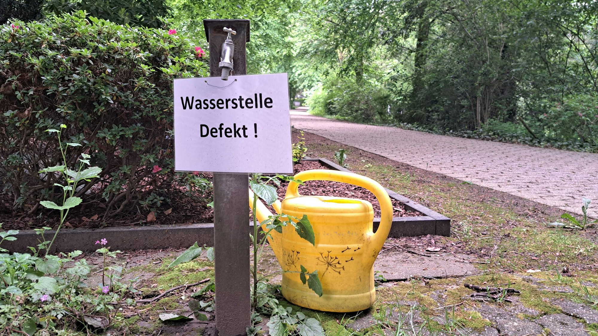 Das Foto zeigt einen Wasserhahn auf dem Wipperfürther Westfriedhof mit der Aufschrift "Wasserstelle defekt", darunter steht eine gelbe Gießkanne.