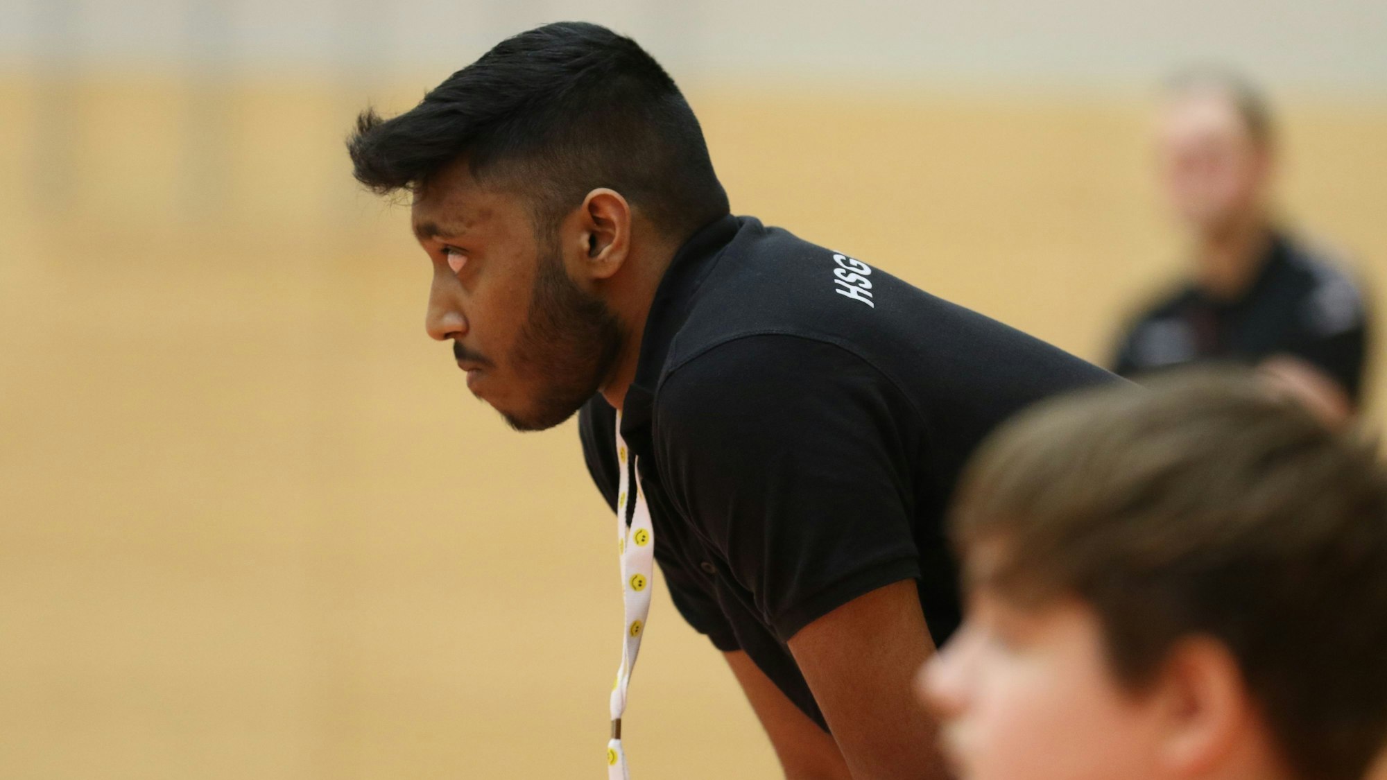 Ein Trainer schaut vornübergebeugt ein Handballspiel. Eine Karte hängt ihm um den Hals.