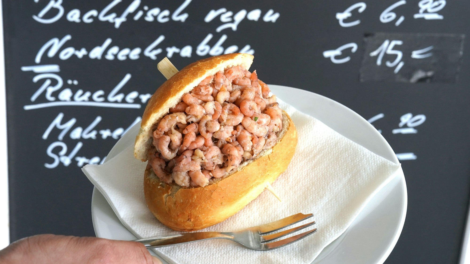 PRODUKTION - 18.04.2024, Hamburg: ILLUSTRATION - Ein Mitarbeiter hält ein Nordseekrabben-Brötchen vor einer Preistafel mit der Aufschrift ·Nordseekrabben - 15,- Euro· an einem Fischbrötchen-Imbiss an den Landungsbrücken hoch.