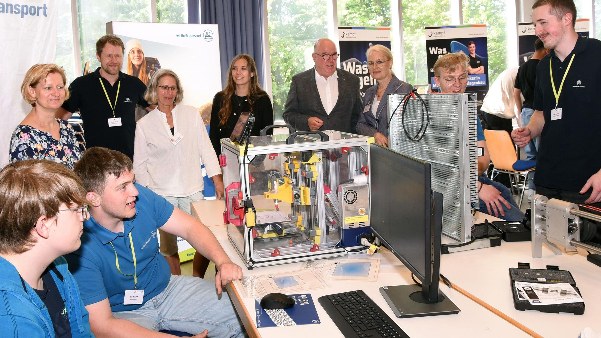 Junge Leute arbeiten an einer technischen Apparatur, Lehrer und Offizielle stehen dahinter.