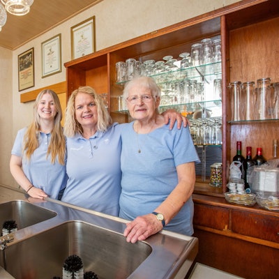 Drei Frauen dreier Generationen stehen in einem Gastraum hinter der Theke nebeneinander.