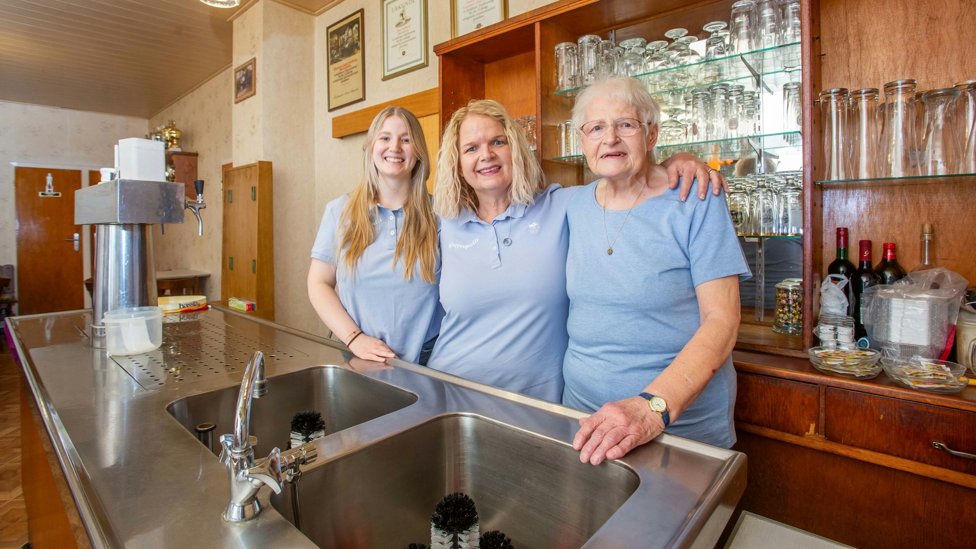 Drei Frauen dreier Generationen stehen in einem Gastraum hinter der Theke nebeneinander.