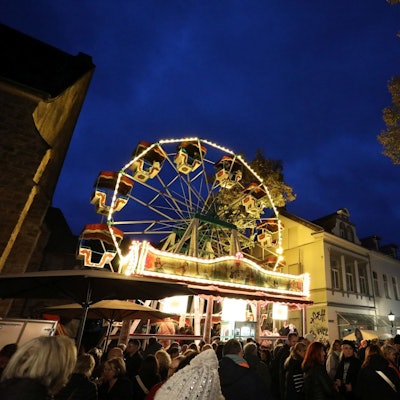 Menschen tummeln sich auf einem Platz, auf dem mehrere Buden und ein beleuchtetes Riesenrad stehen.