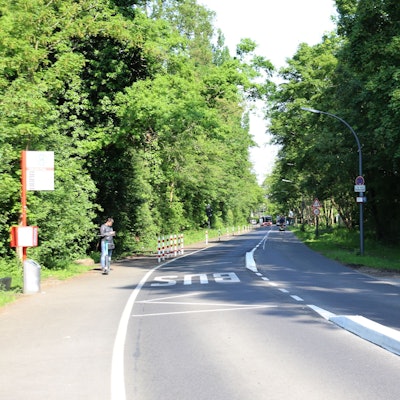 Blick auf den Abschnitt Zeisbuschweg zwischen einer Bushaltestelle und dem Verbindungsweg zur Straße Am Puffelskooche (im Bild rechts). Die Straße ist umsäumt von grünen Bäumen. Foto von Uwe Schäfer
