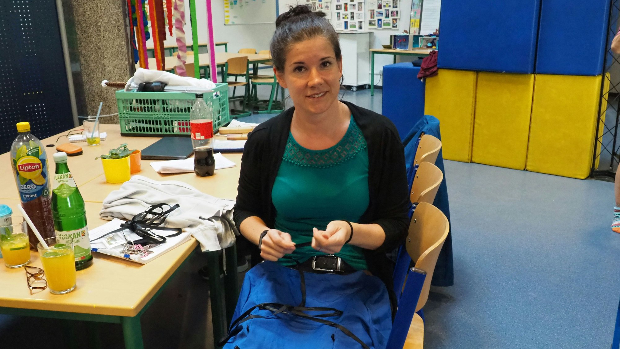 Susanna Janssen hat ihr Kostüm auf dem Schoß und arbeitet daran, im Hintergrund sind Requisiten zu sehen.