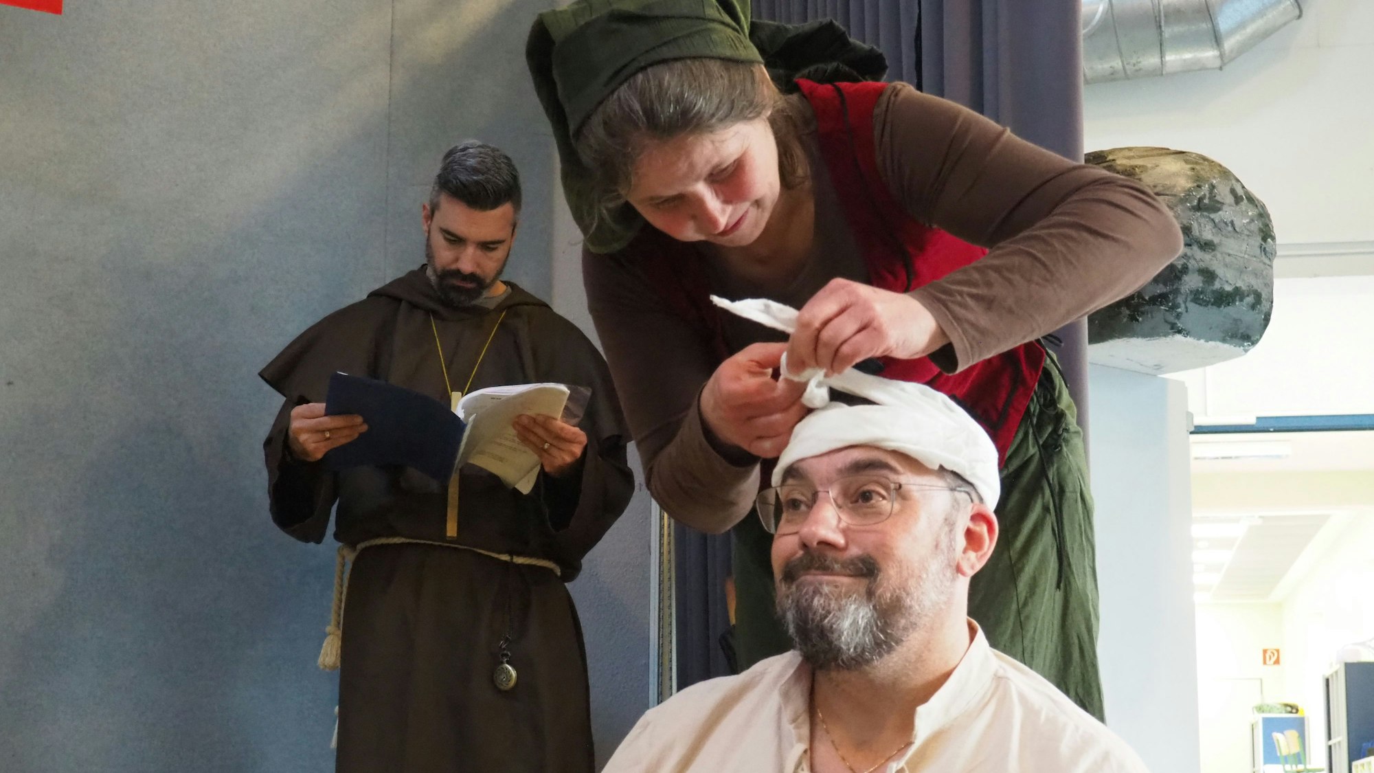 Eine Frau verbindet einem Mann den Kopf, im Hintergrund steht ein Geistlicher. e