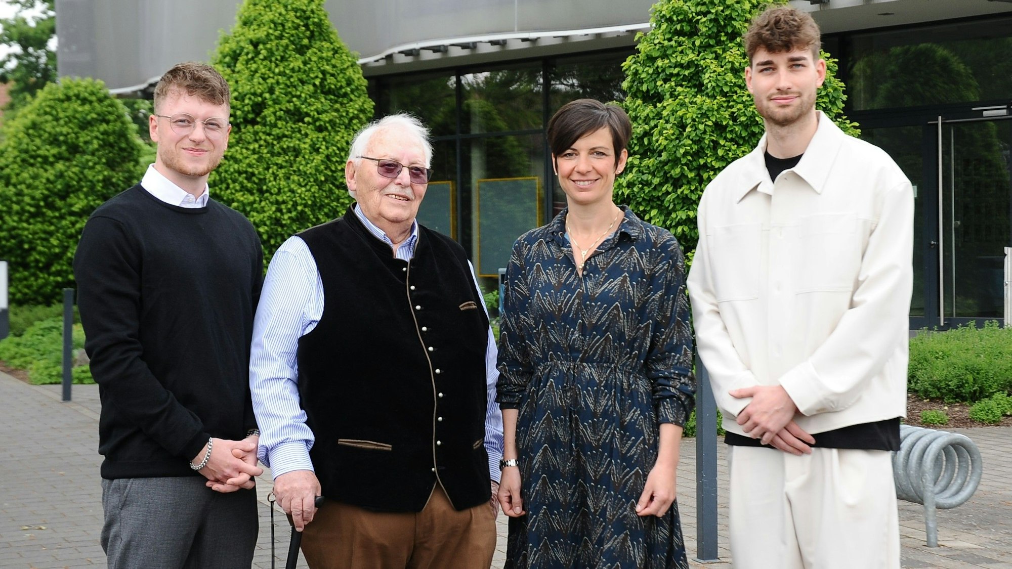 Drei Männer und eine Frau stehen vor einem grauen Gebäude, im Hintergrund sind Büsche zu sehen.