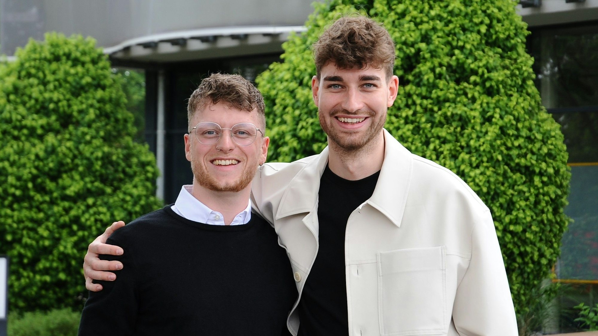 Zwei Männer stehen vor einem grauen Gebäude, im Hintergrund sind Büsche zu sehen.