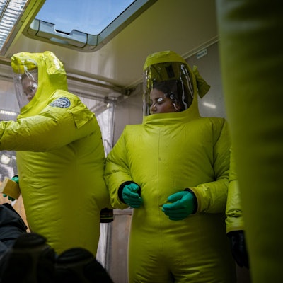 Auf dem Foto sind Feuerwehrleute in Schutzanzügen zu sehen, die einen Kollegen, der einen Patienten mimt, behandeln.