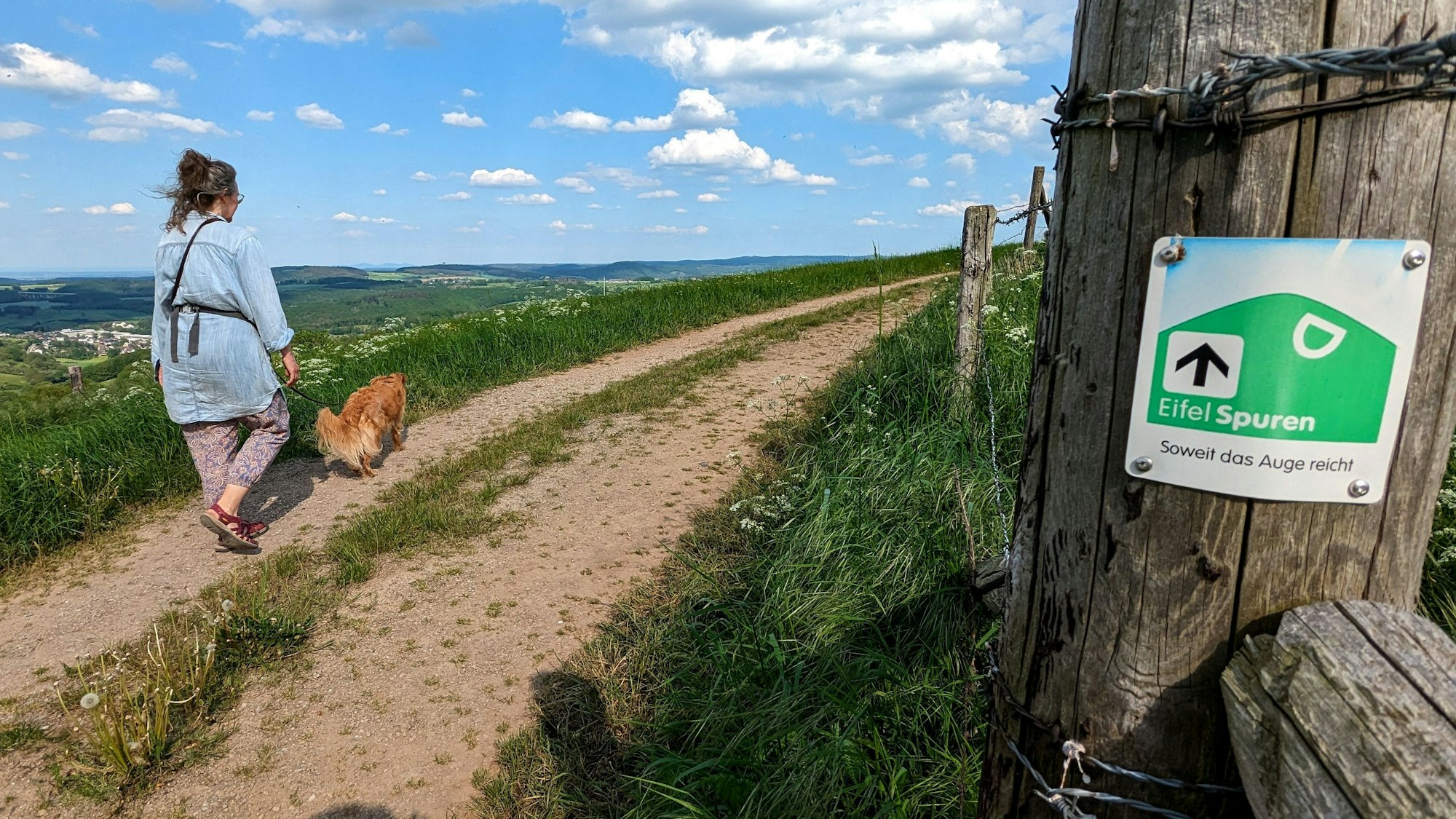 Eine Spaziergängerin und ihr Hund auf der Eifelspur „Soweit das Auge reicht“. Rechts an einem Zaunpfahl ist eine Wegmarkierung mit dem Titel des Weges angebracht.