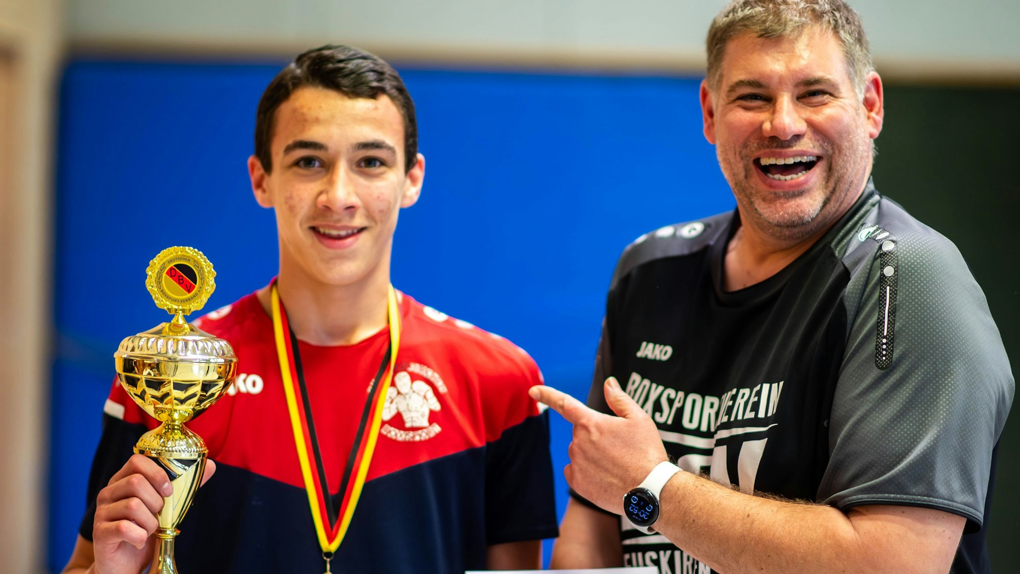 Das Bild zeigt den jungen Boxer, der den Pokal in die Kamera hält. Der Vorsitzende des Vereins lacht und zeigt mit dem Finger stolz auf das Boxtalent.
