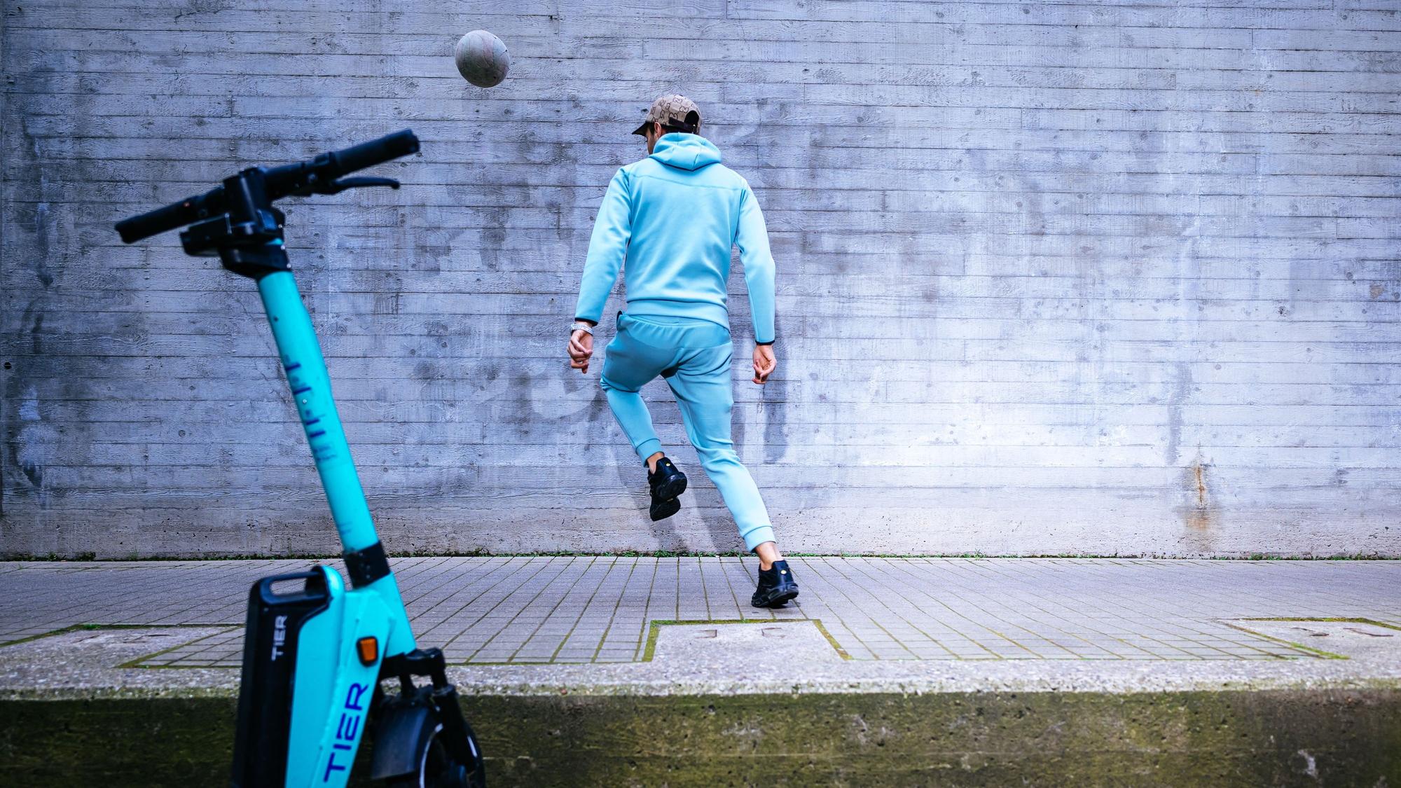 In Bewegung: Ein junger Mann kickt vor einem E-Scooter einen Ball an eine Hauswand.
