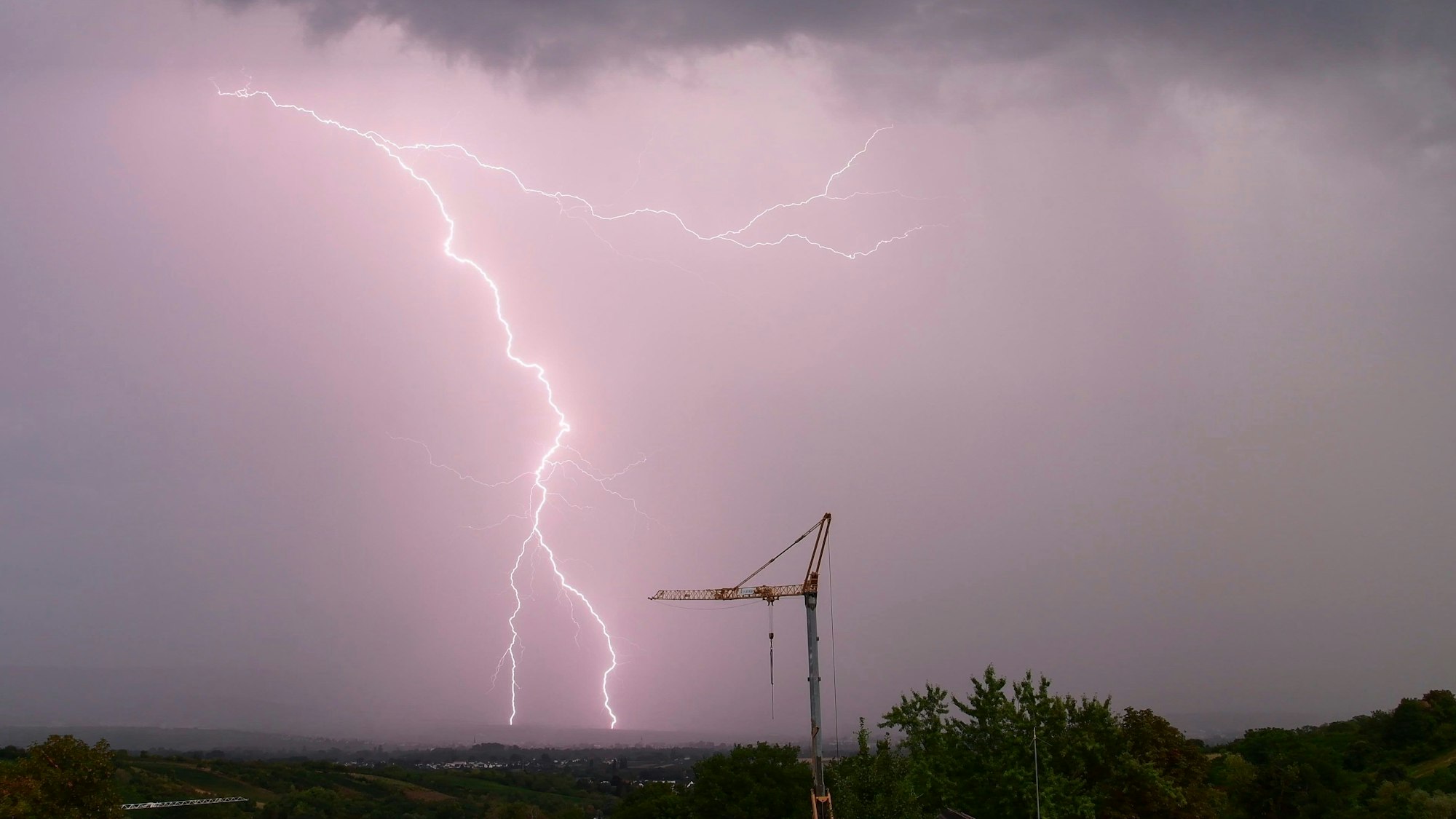 Ein gewaltiger Blitz schlägt am vom Unwetter lila gefärbten Himmel in der Nähe eines Baukrans ein. Schwere Unwetter werden in der Nacht zu Freitag in Köln und der Region erwartet. (Archivbild)