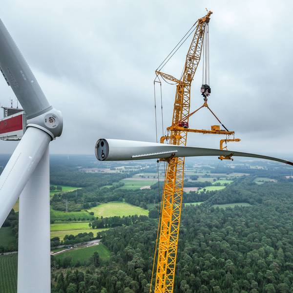 Bau einer Windenergieanlage. Ein mobiler Großkran hebt ein Rotorblatt zum Maschinenhaus auf den Turm.