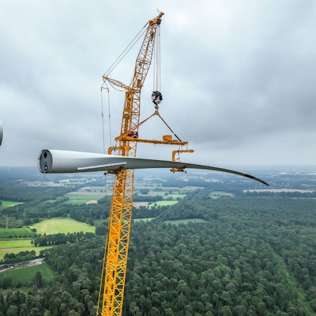 Bau einer Windenergieanlage. Ein mobiler Großkran hebt ein Rotorblatt zum Maschinenhaus auf den Turm.