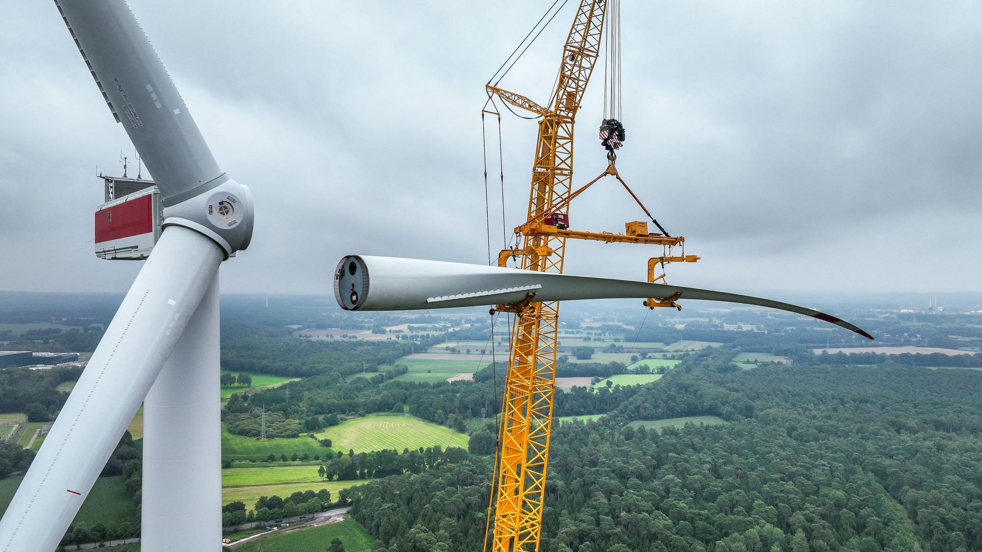 Bau einer Windenergieanlage. Ein mobiler Großkran hebt ein Rotorblatt zum Maschinenhaus auf den Turm.
