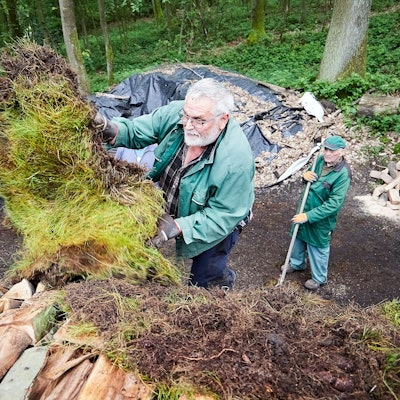 Gerd Linden bedeckt die gestapelten Scheite mit Grassoden, ein Helfer schaut von unten zu.