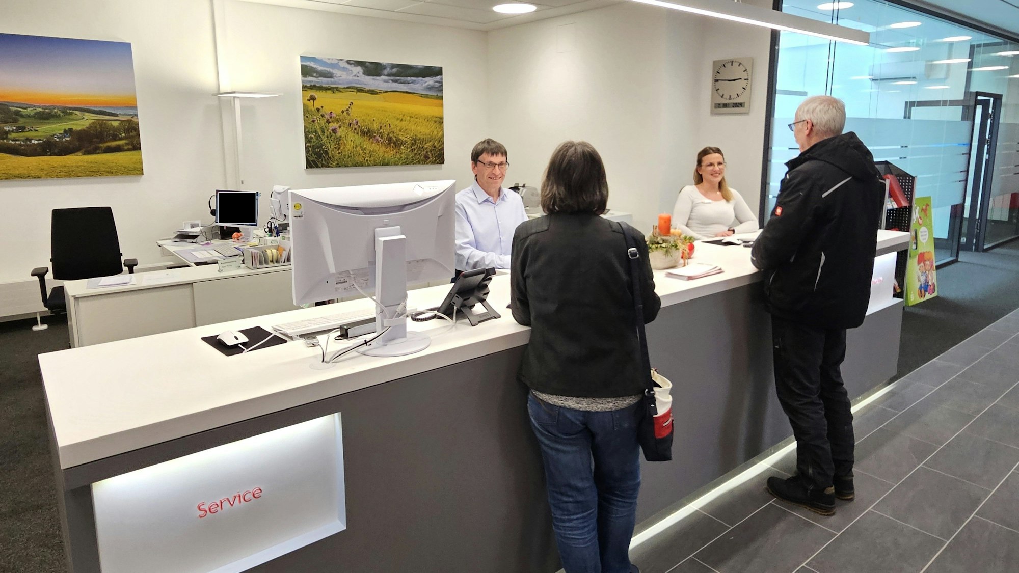 Die Servicemitarbeiter Egon Ronig und Denise Maletzki stehen am Schalter und bedienen zwei Kunden.
