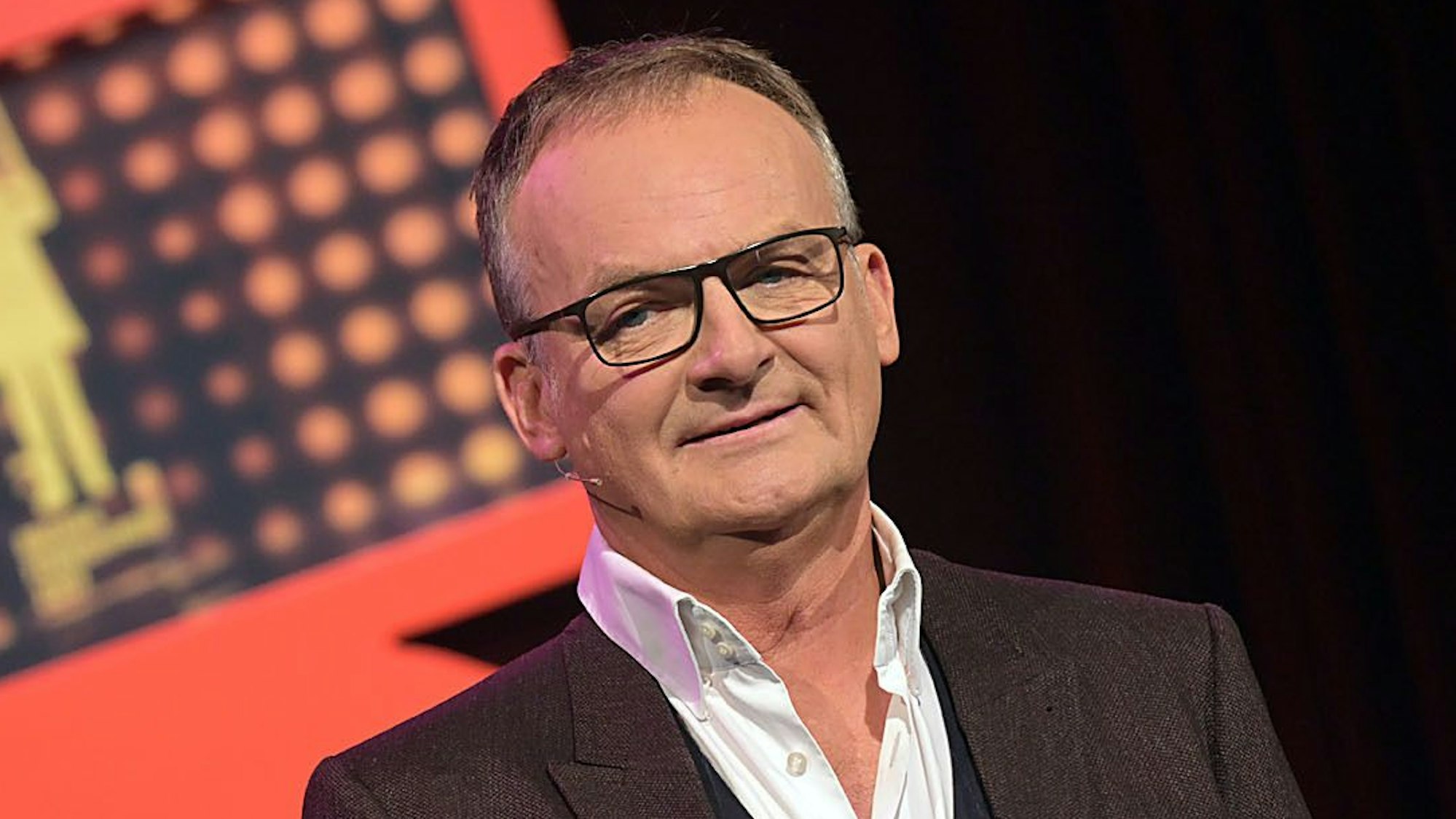 Moderator Frank Plasberg, bekannt durch die ARD-Talkshow „Hart aber fair“, steht in Anzug und Hemd auf der Bühne und blickt in die Kamera.
