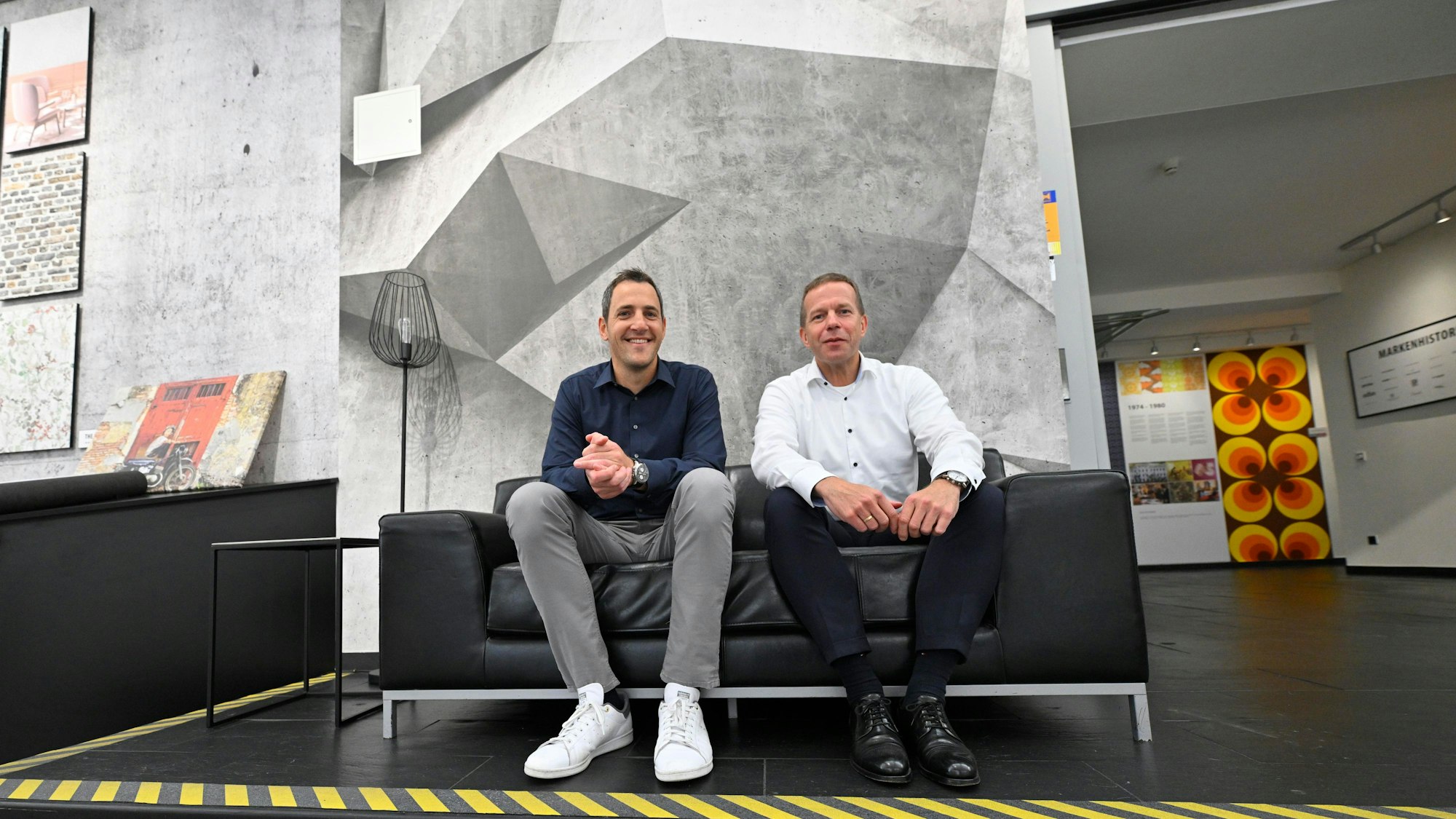 Zwei Herren sitzen auf einem Sofa, dahinter Mustertapeten.