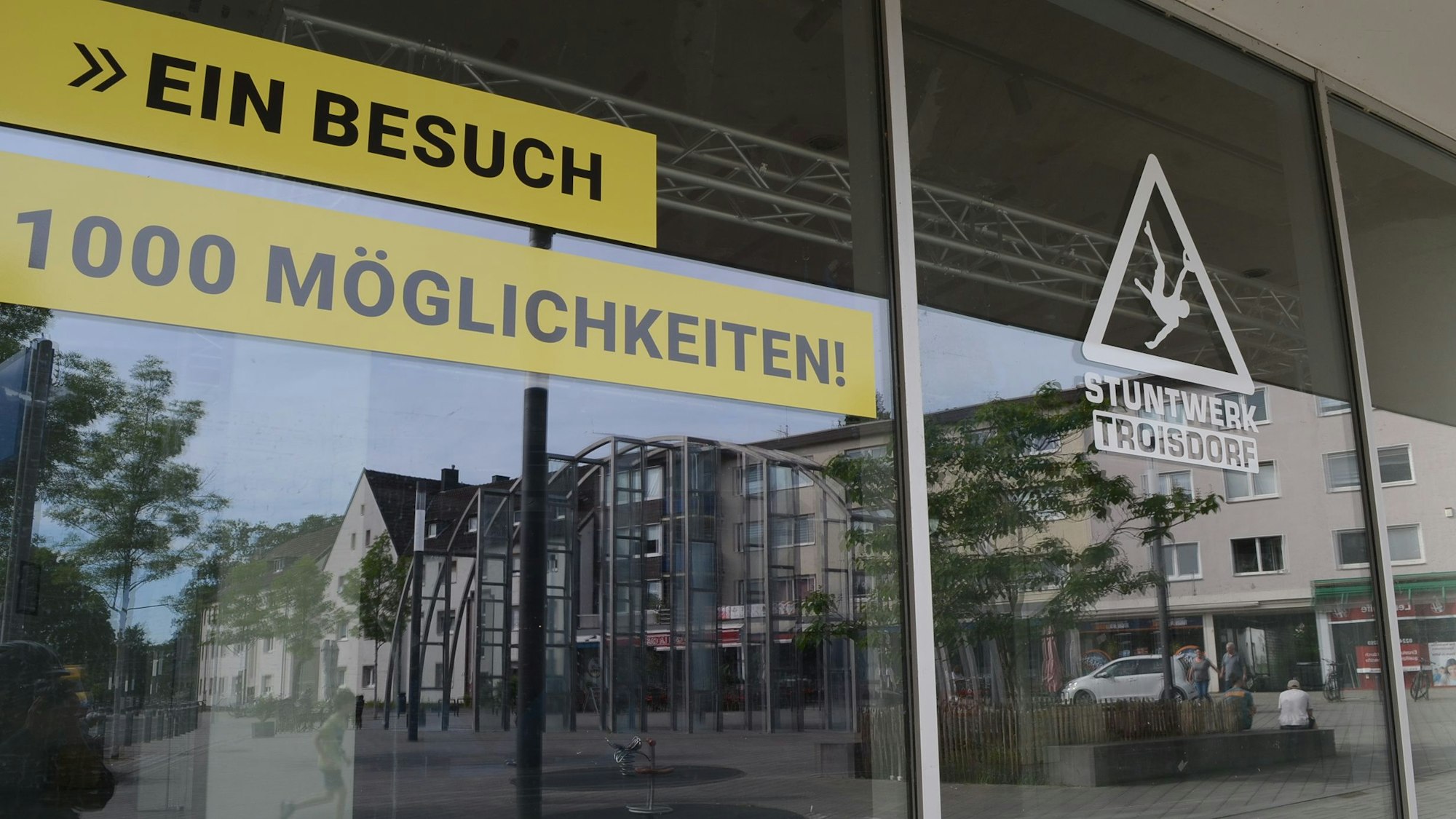 Eine Schaufensterscheibe mit Werbeaufschrift: „Ein Besuch 1000 Möglichkeiten;“ rechts daneben steht „Stuntwerk Troisdorf“. Im Fenster spiegeln sich die Häuser von gegenüber.