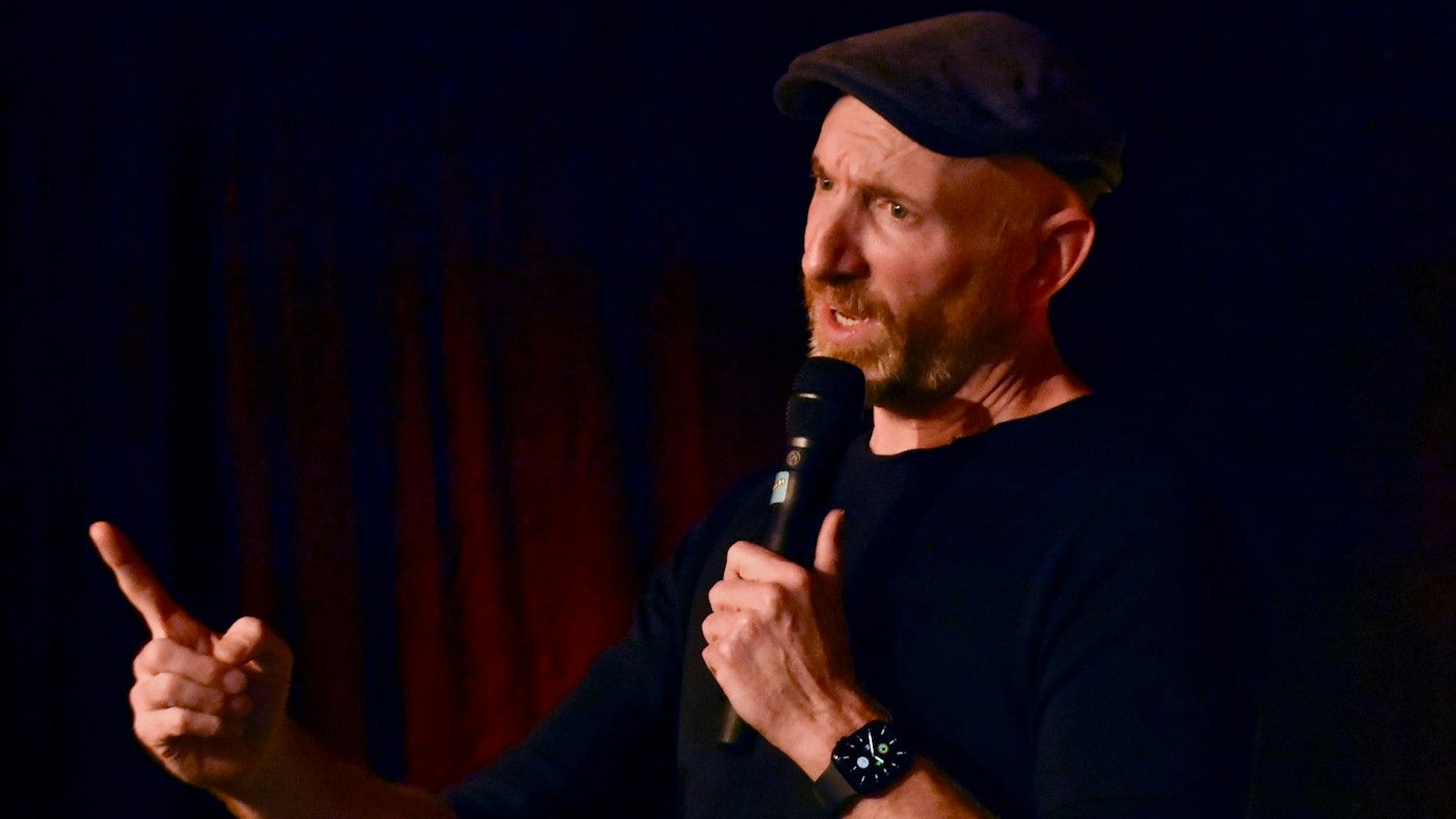 Ein Mann im schwarzen T-Shirt und einer schwarzen Schiebermütze ; er hält ein Mikrophon in der Hand.