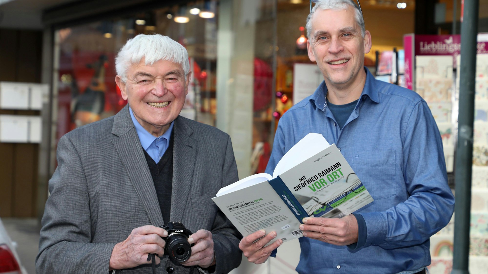 Siegfried Raimann steht mit einer Kamera in der Hand neben Verleger und Buchhändler Alexander Bücken, der sein Buch „Mit Siegfried Raimann vor Ort“ in den Händen hält.