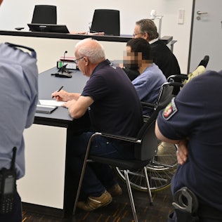 Der Angeklagte sitzt in einem Gerichtssaal neben seinem Verteidiger in einem Rollstuhl.