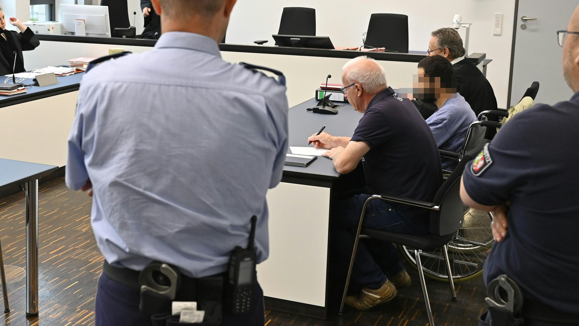 Der Angeklagte sitzt in einem Gerichtssaal neben seinem Verteidiger in einem Rollstuhl.